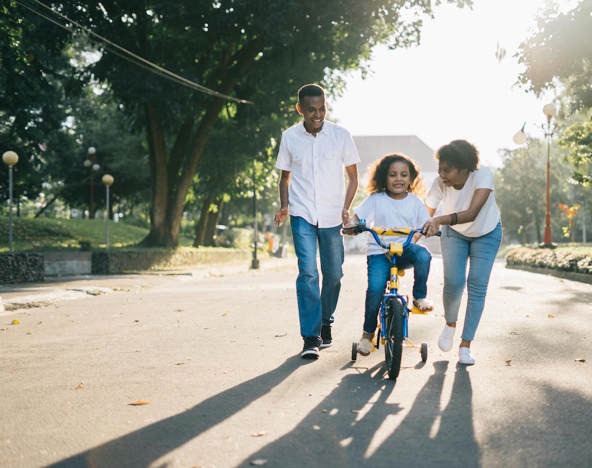 Young husband and wife chase child on bicycle with training wheels teaching them how to ride